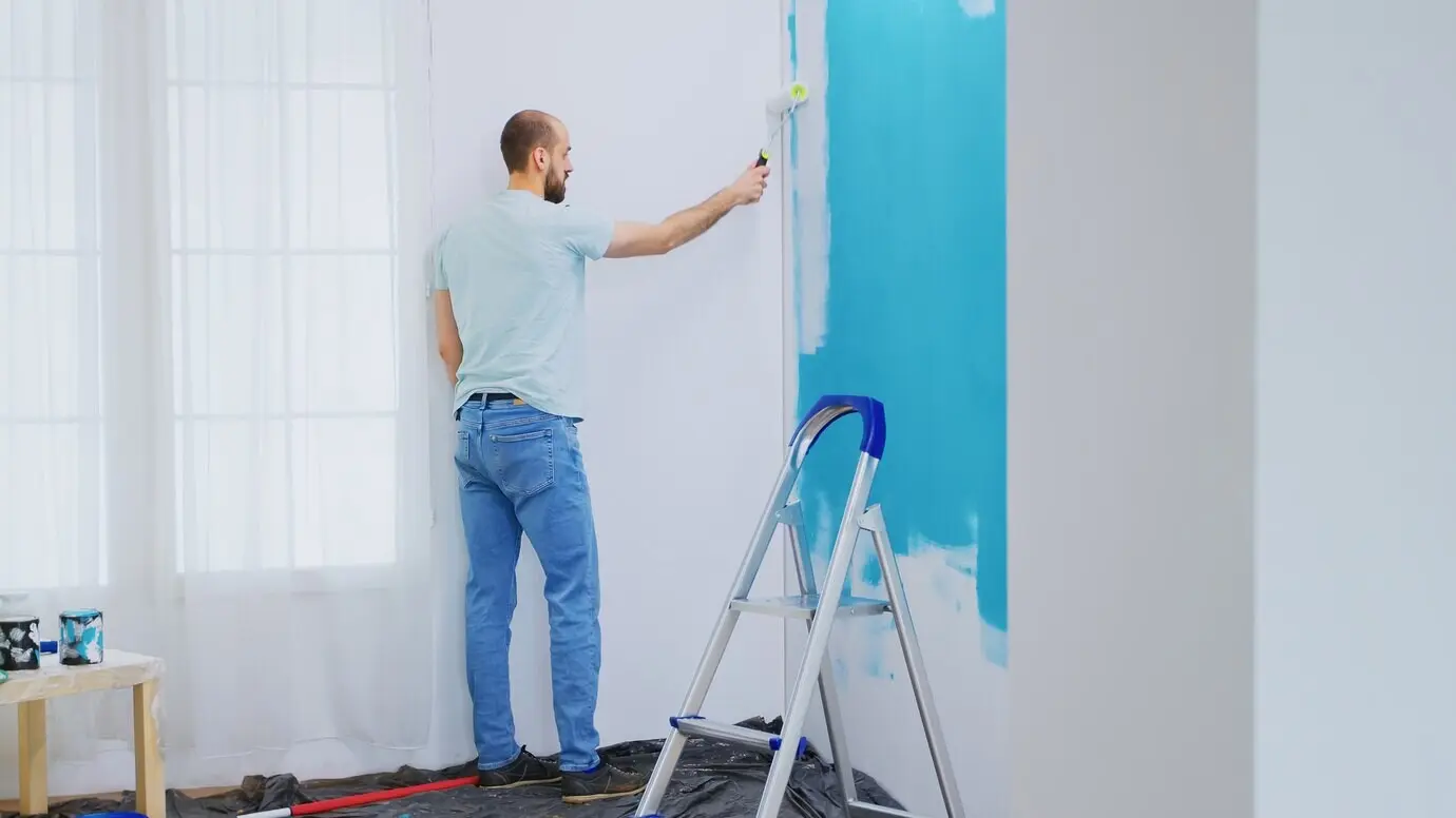 Eine blaue Wand wird während der Wohnungsrenovierung mit weißer Farbe und einer Farbrolle gestrichen. Ein Handwerker renoviert. Wohnungsumgestaltung und Bauarbeiten im Haus im Rahmen der Renovierung und Verbesserung. Reparatur und Dekoration.